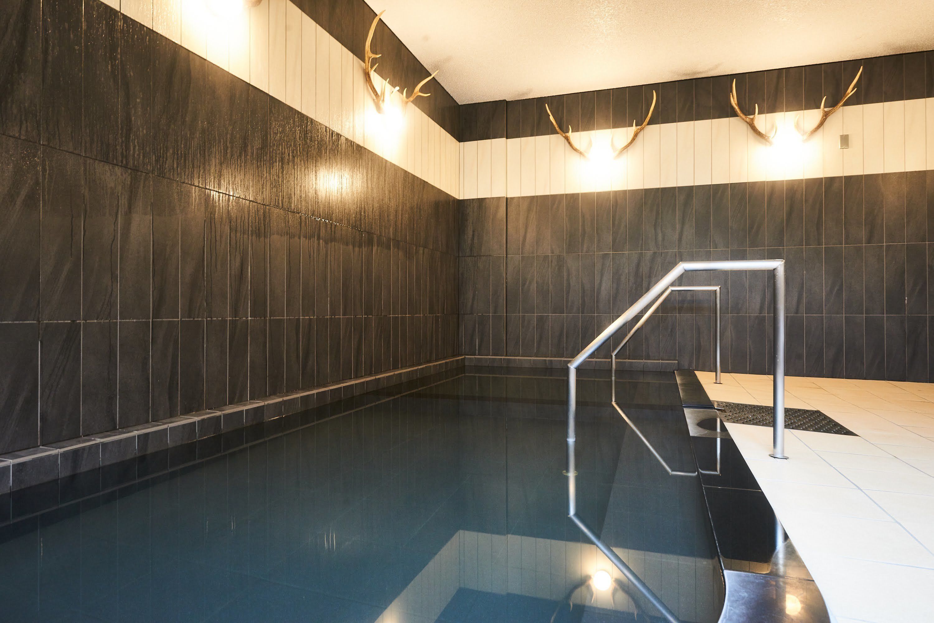 Un área de piscina interior moderna con paredes de azulejos oscuros, una barandilla de vidrio transparente y adornos de cuernos en la pared.