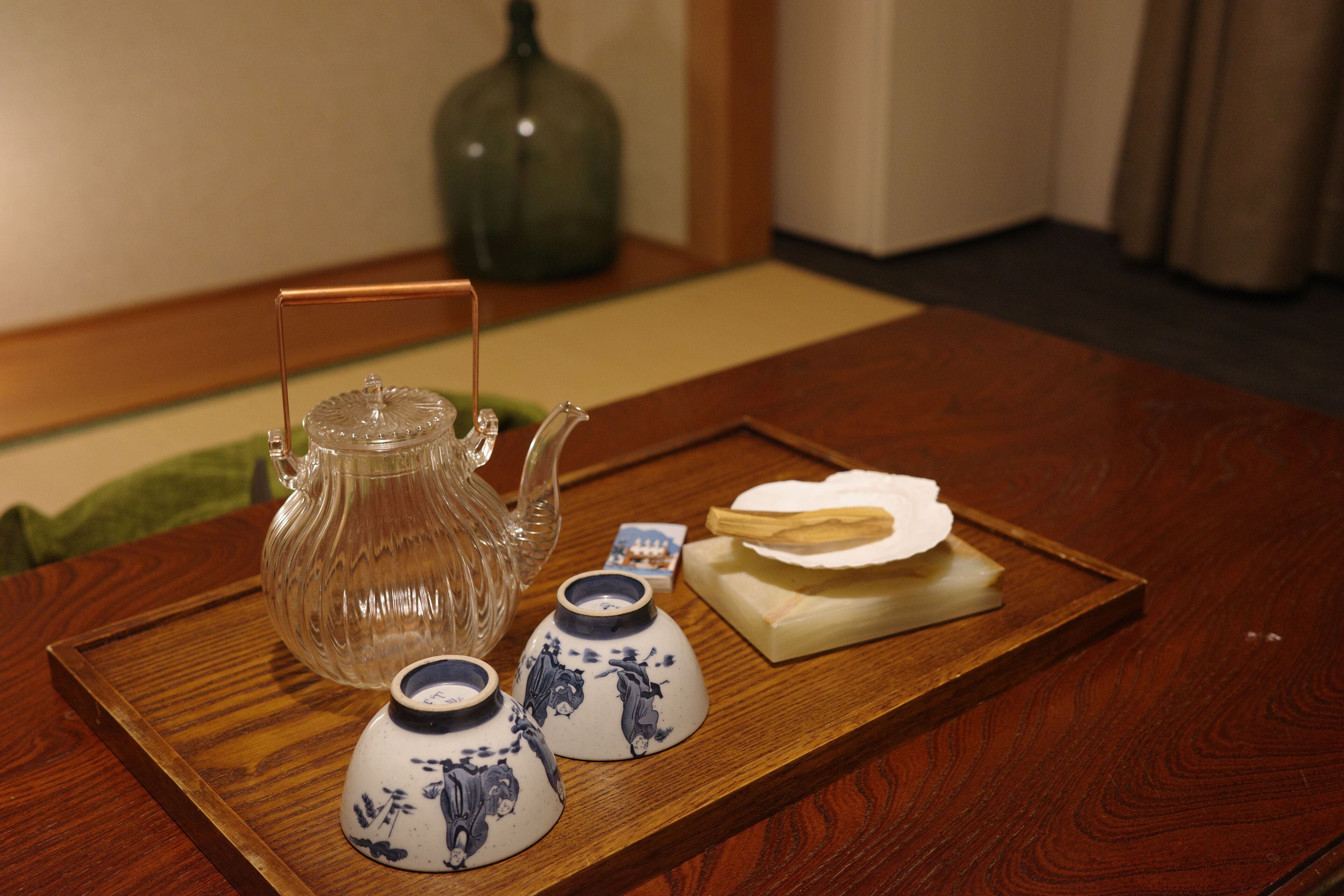A wooden tray with a glass teapot, two ceramic cups, and a stack of white plates on a table.