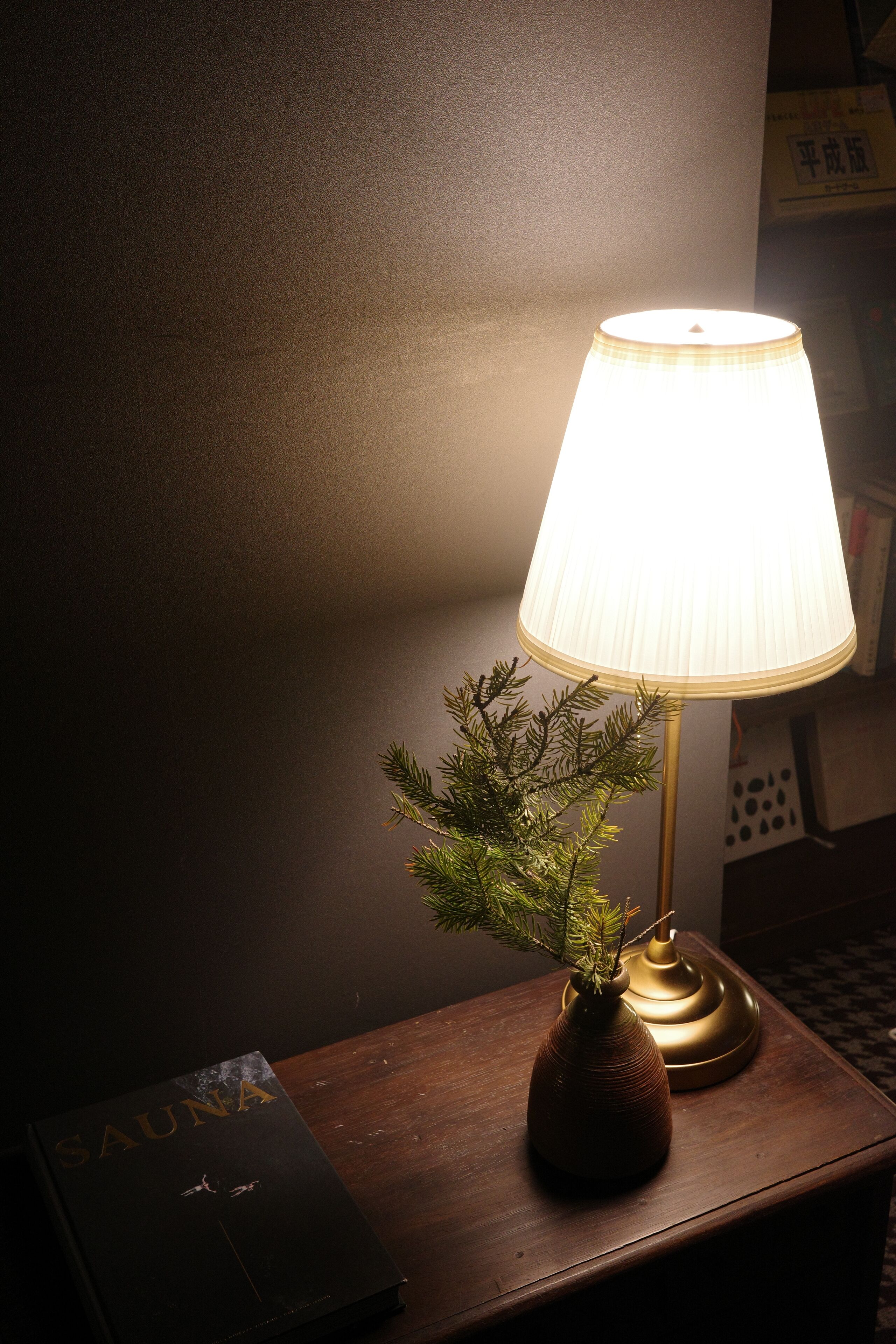 A wooden nightstand with a lamp, a book titled "SAUNA," and a small vase with a plant.
