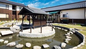 A modern building with a covered outdoor seating area, a circular stone water feature, and a well-maintained garden. - Racine Home Harie (Takashima)