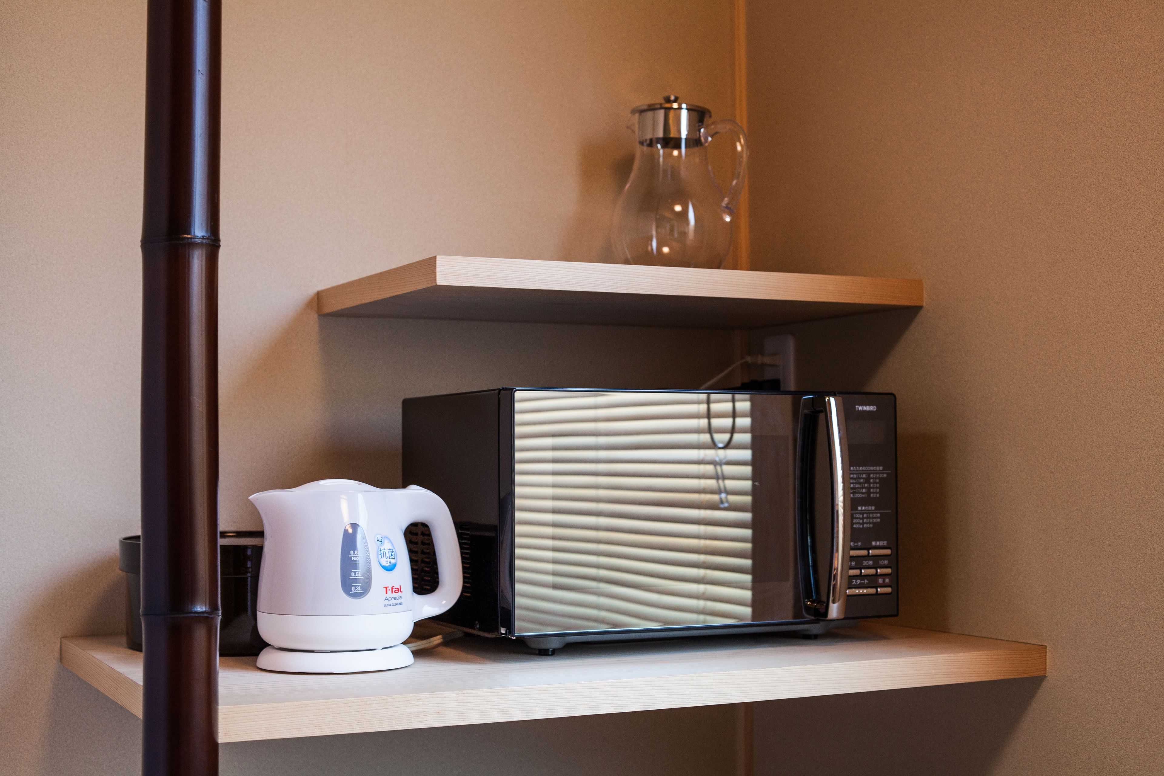 A kitchen area with a microwave, a kettle, and a glass pitcher on a shelf.