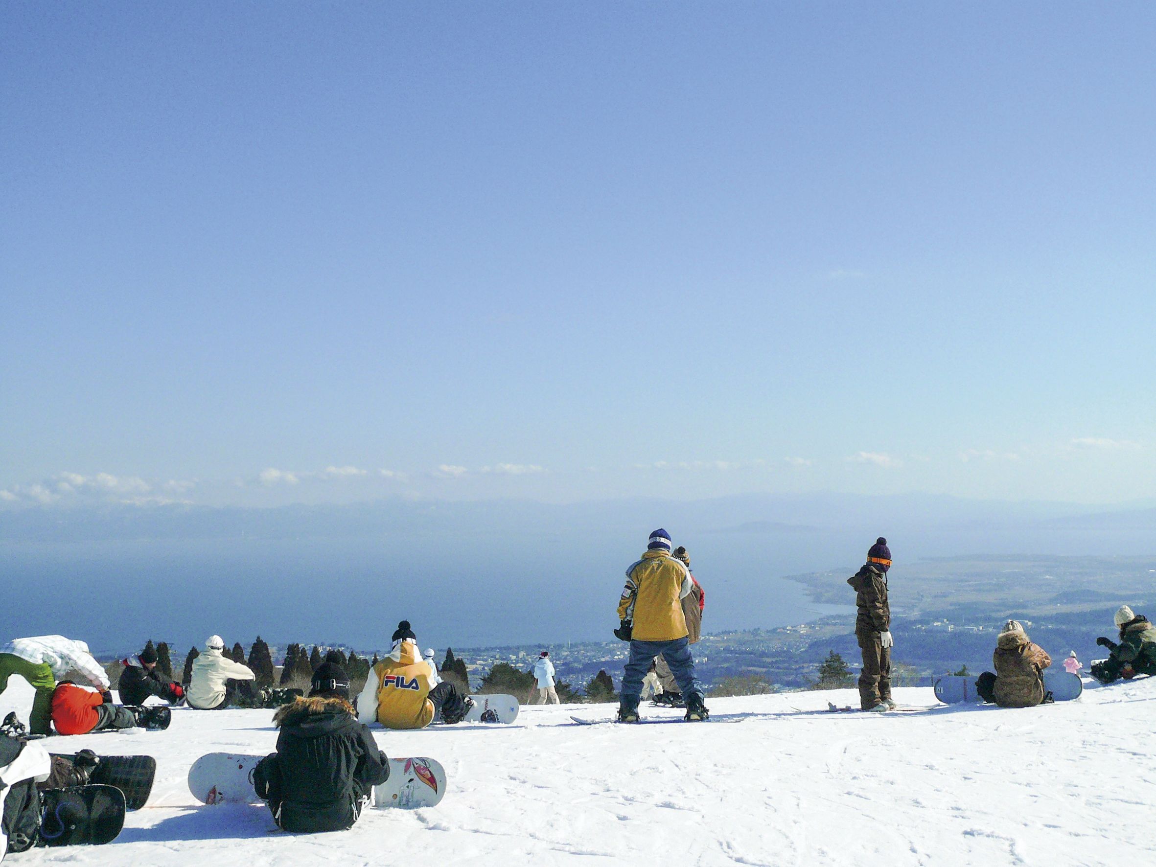 A group of people on a snowy mountain with snowboards, sitting and standing on a clear day.