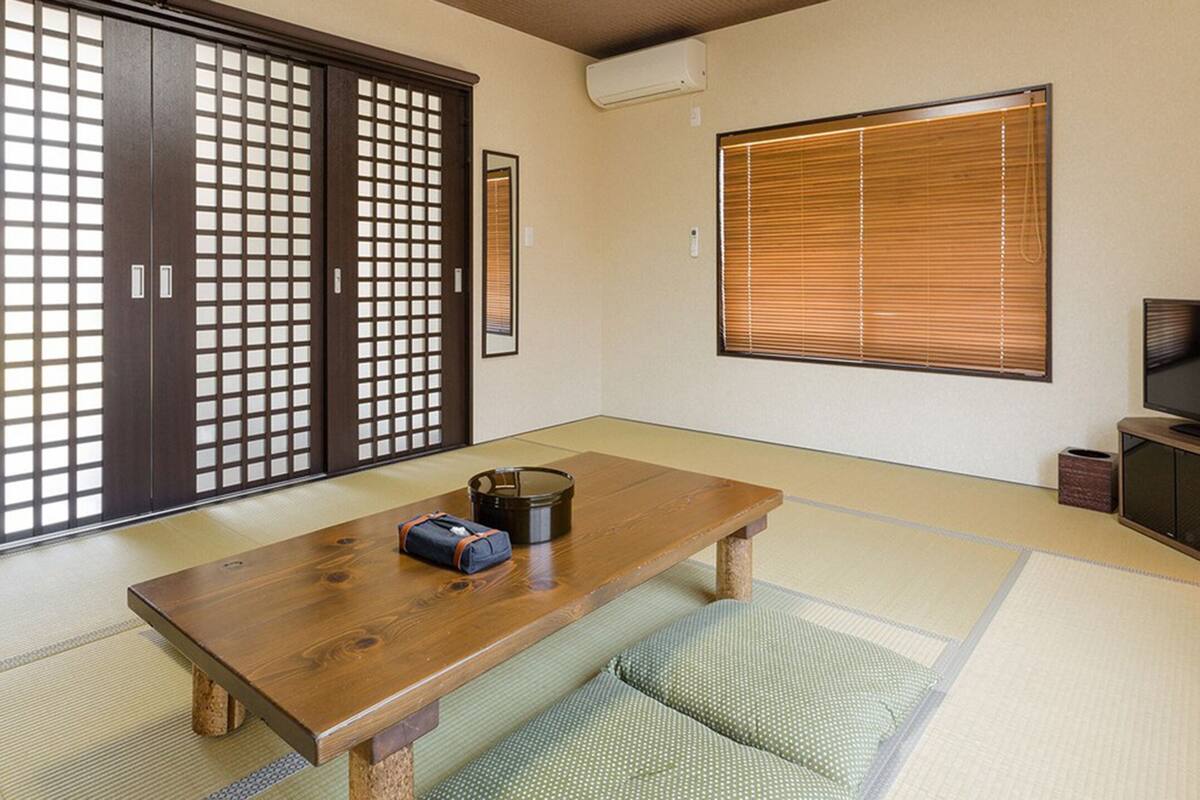 A traditional Japanese room with tatami flooring, a wooden table, a television, sliding doors, and a window with blinds.