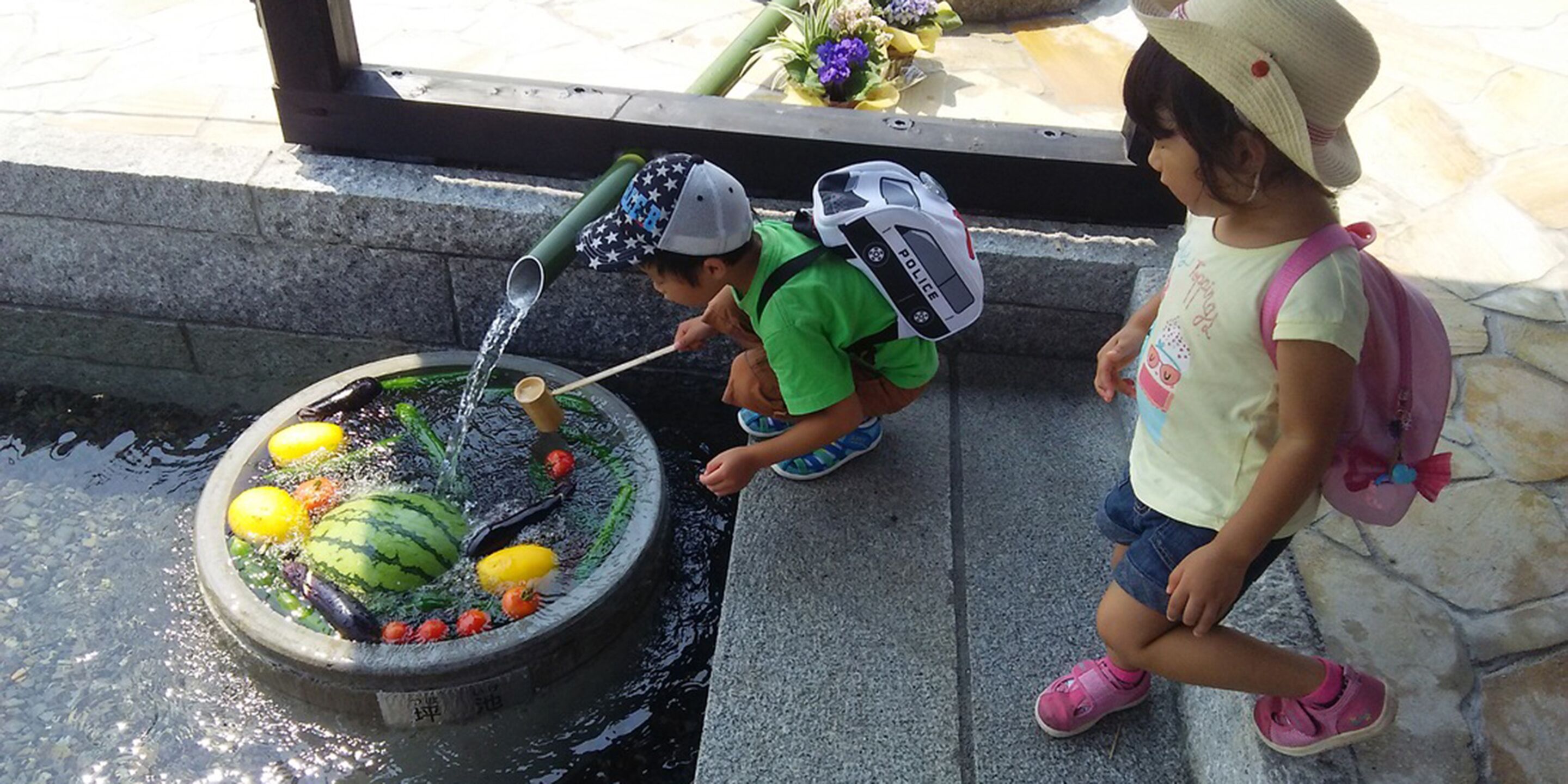 A child playing with water in a fountain, another child observing, and a police officer nearby.