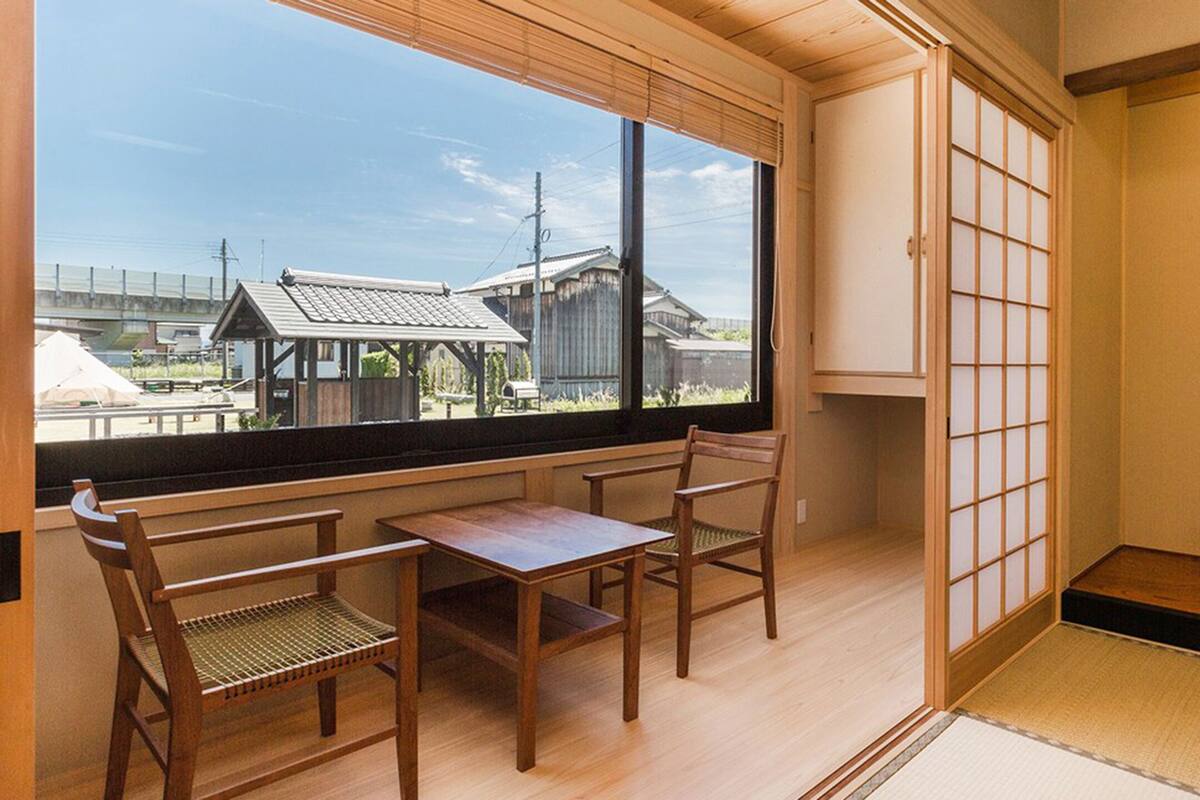 A traditional Japanese room with tatami flooring, a low table with two chairs, a shoji screen, and a bonsai tree.