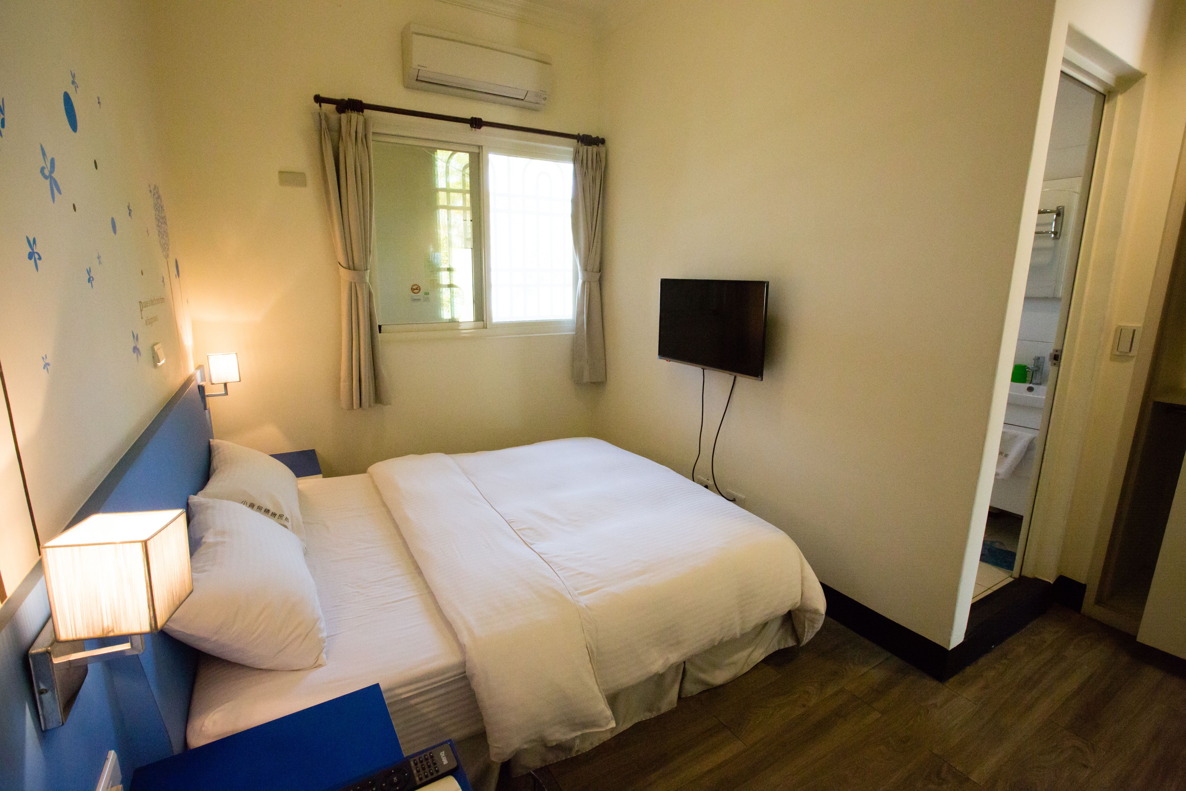 A hotel room with a bed, a TV, a window with curtains, a bedside lamp, and a wall-mounted air conditioner.