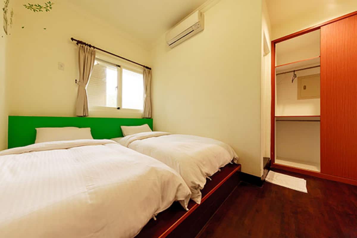 A bedroom with two beds, a green headboard, a window with curtains, an air conditioning unit, and a wooden wardrobe.