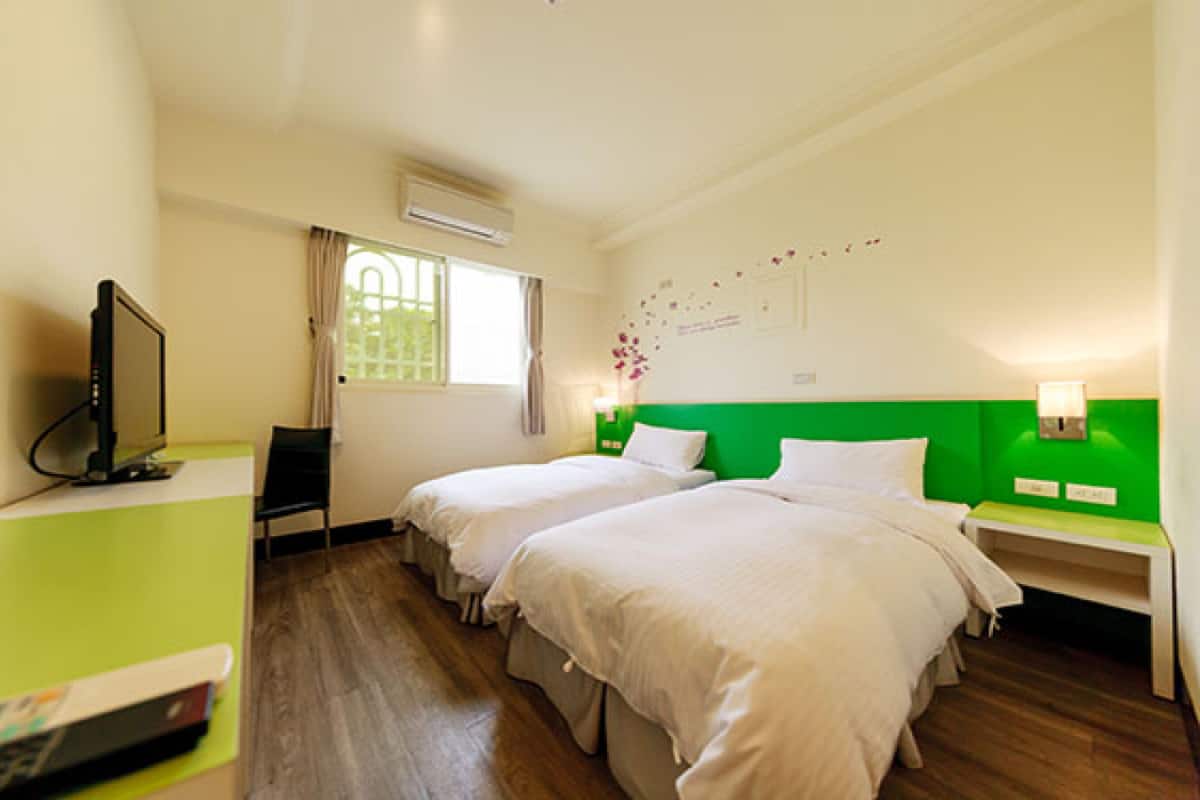 A hotel room with two beds, a TV, a desk with a computer, and a window with curtains.