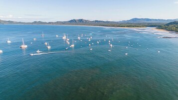 Vista aérea de una zona costera con numerosos barcos anclados en el agua, rodeada por un paisaje montañoso.