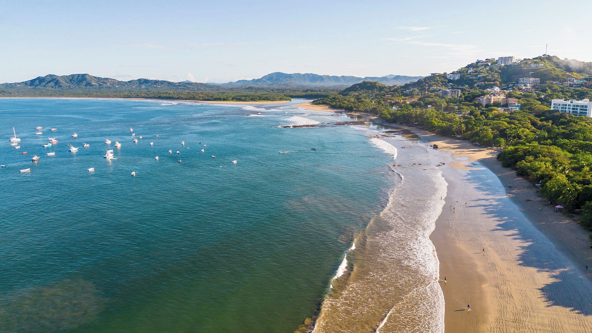 A coastal beach with clear waters, boats, and a hilly landscape with buildings.