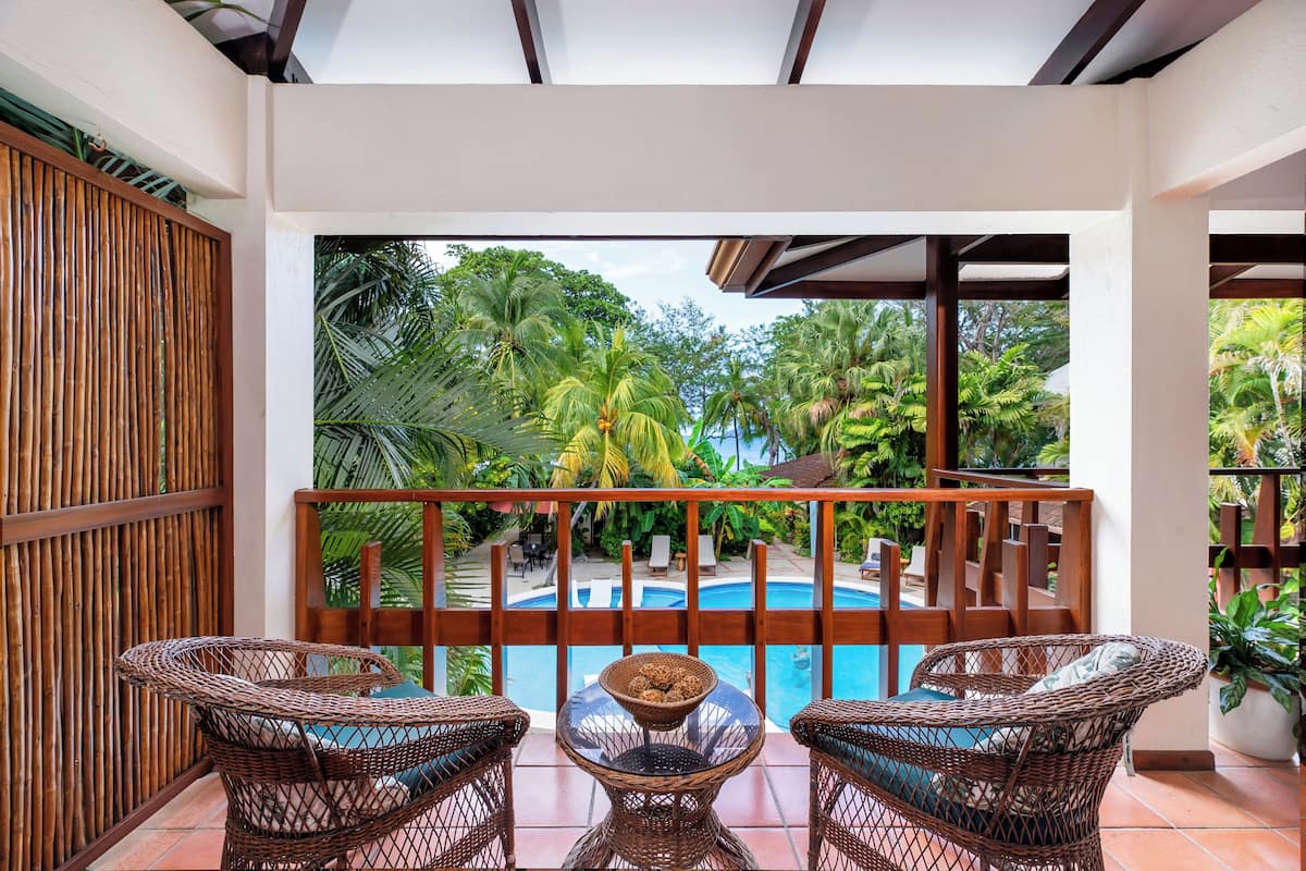 Ein Balkon mit Korbmöbeln, einem Glastisch und Blick auf einen Pool, der von tropischer Vegetation umgeben ist.