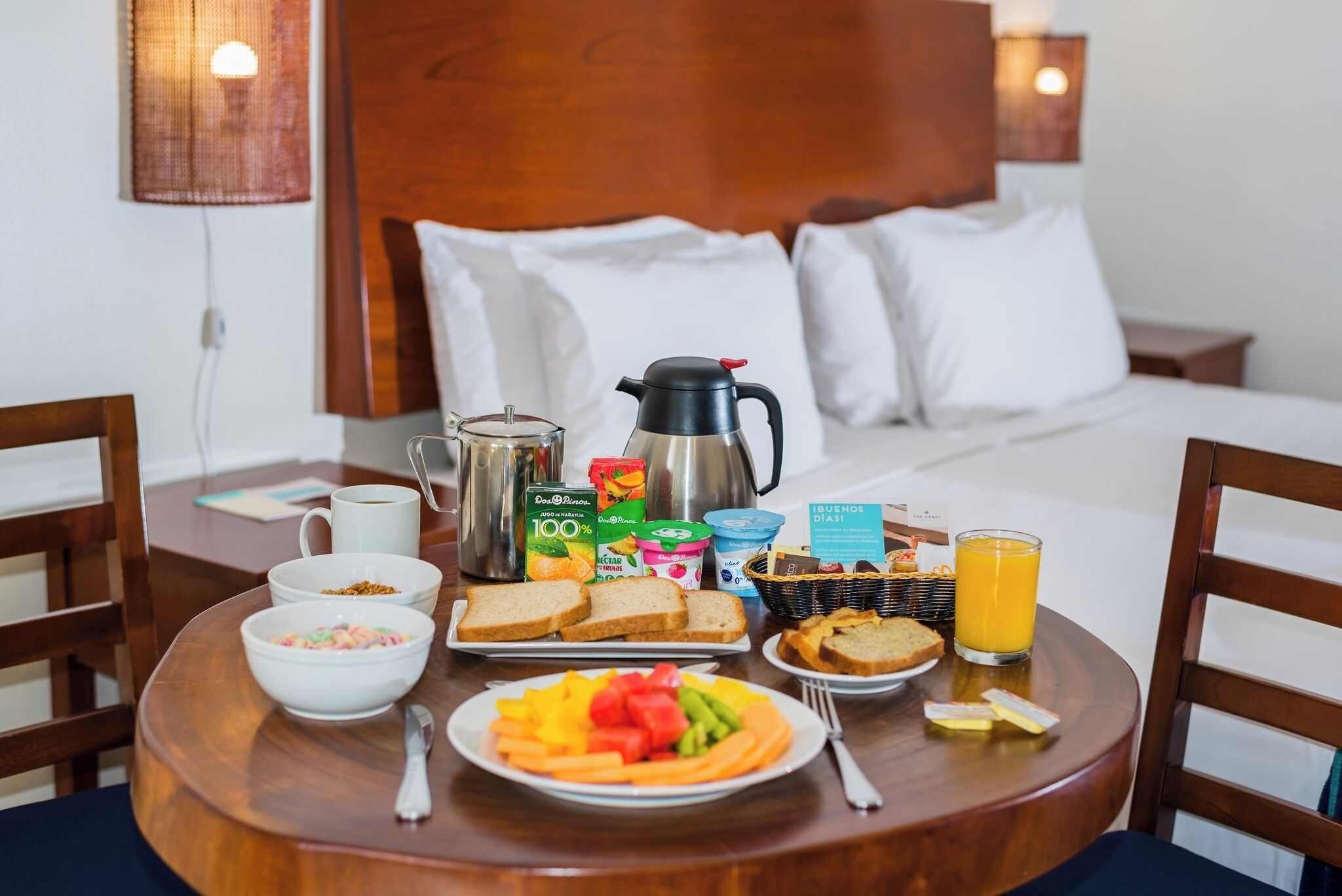 Una mesa de madera con un desayuno que incluye una cafetera, cereal, tostadas, fruta, yogur y jugo.