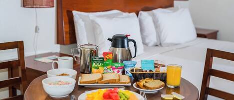 Una mesa de madera con un desayuno que incluye una cafetera, cereal, tostadas, fruta, yogur y jugo.
