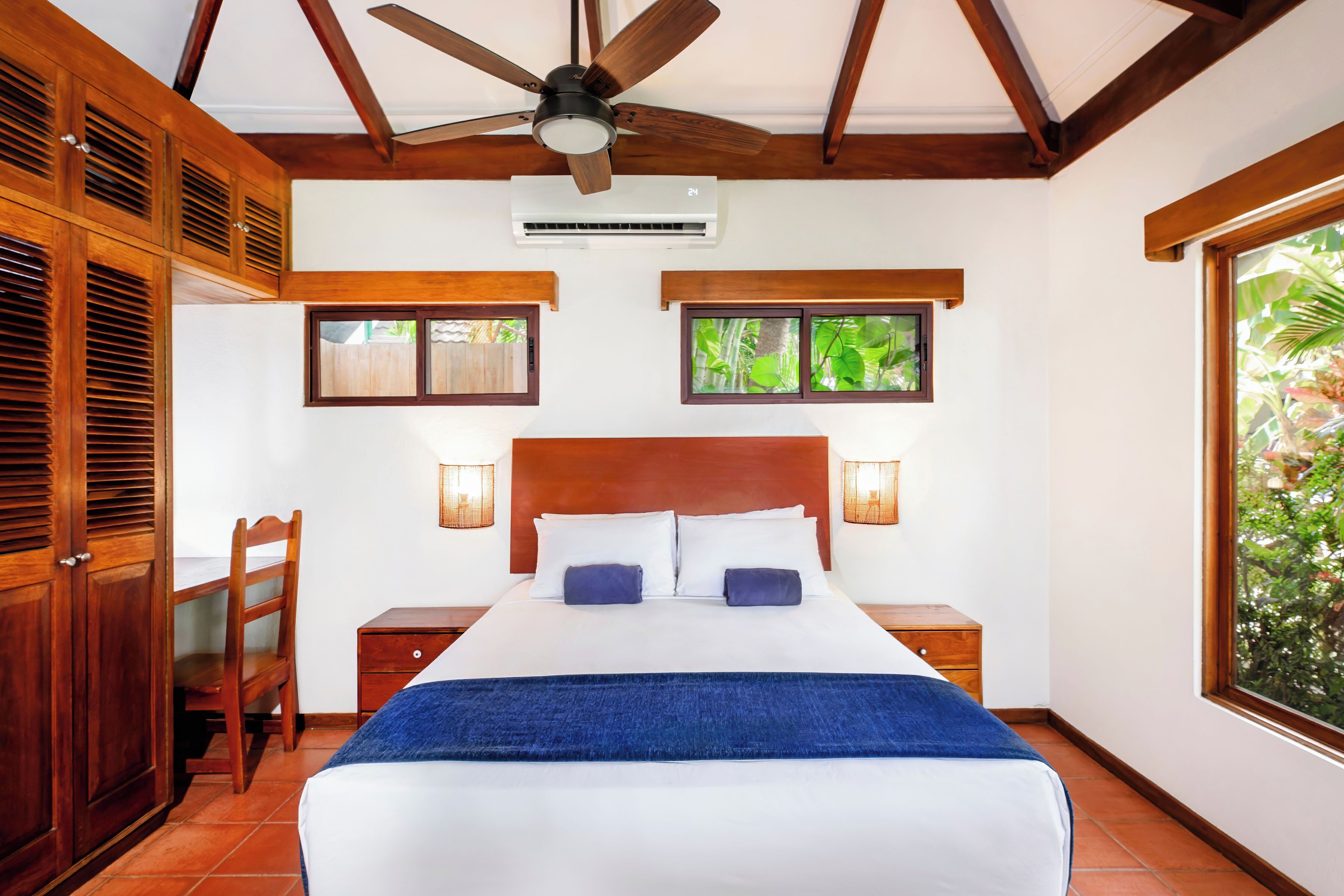 A bedroom with a bed, wooden headboard, two bedside tables with lamps, a ceiling fan, and windows with views of greenery.