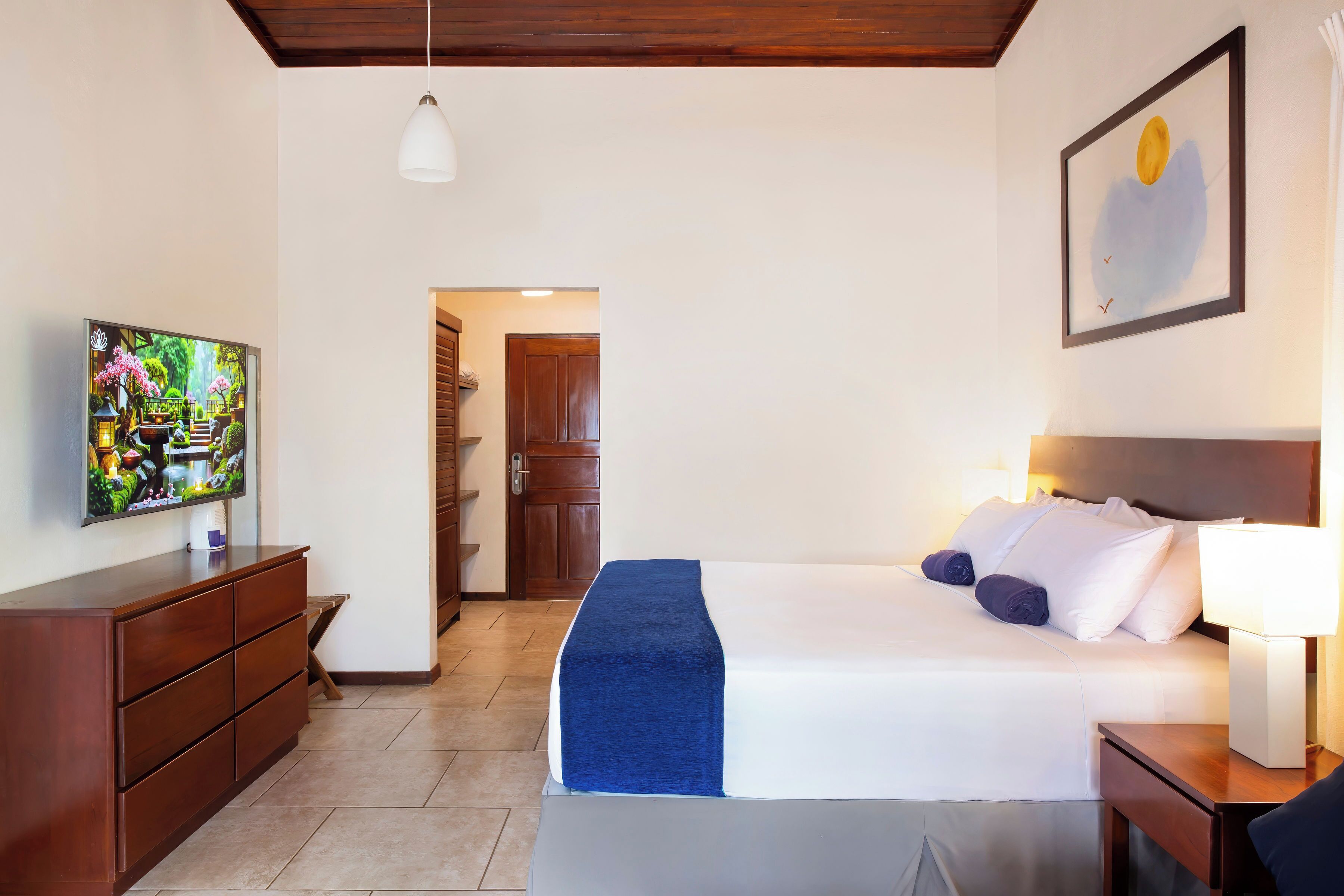 Une chambre d’hôtel avec un lit, un téléviseur posé sur une commode en bois, un tableau accroché au mur et une porte donnant sur une autre pièce.