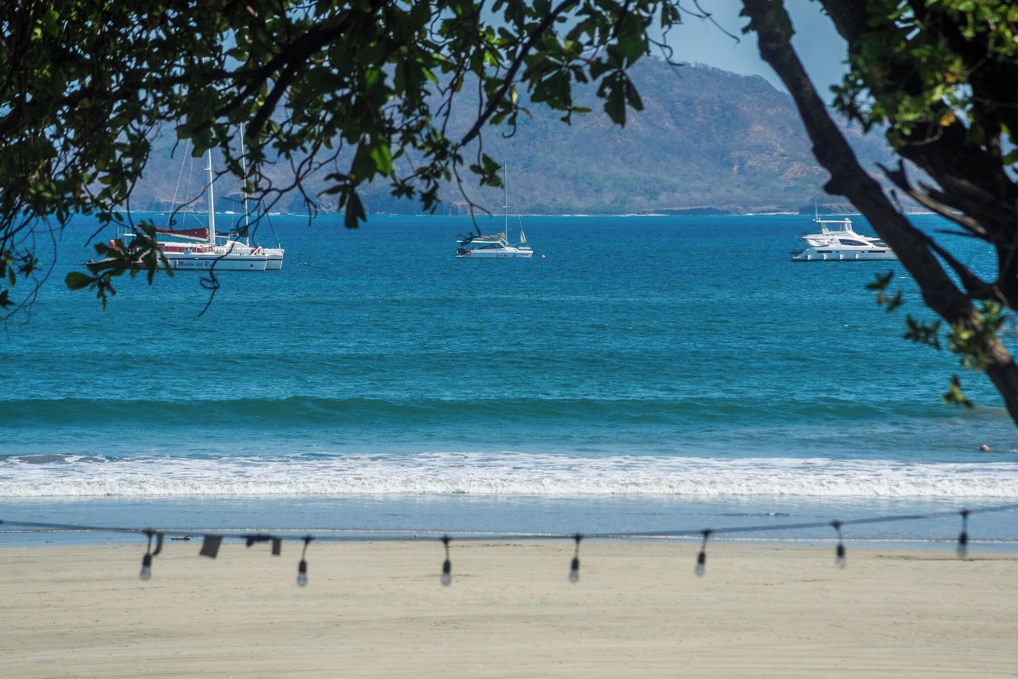Una playa con tres veleros en el agua, una orilla arenosa y una montaña al fondo.