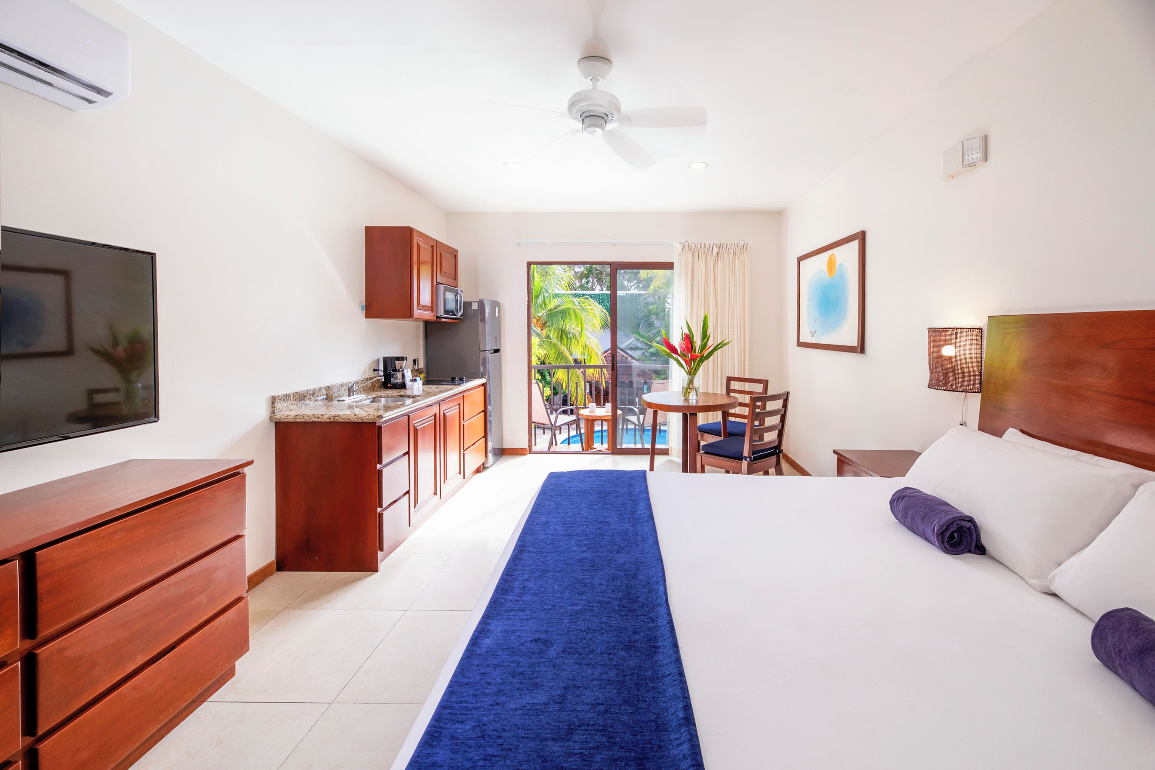 Une chambre d’hôtel équipée d’un lit, d’un bureau, d’une kitchenette et d’un balcon avec vue sur la verdure.