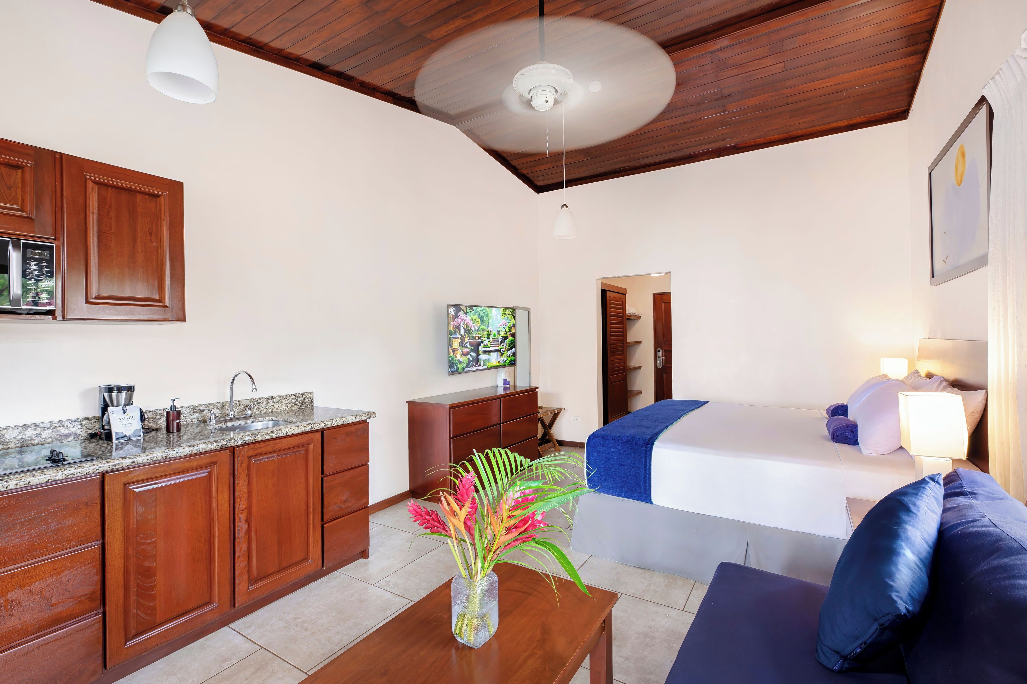 A compact room with a kitchenette, a bed with a blue blanket, a small table with a vase of flowers, and a ceiling fan.