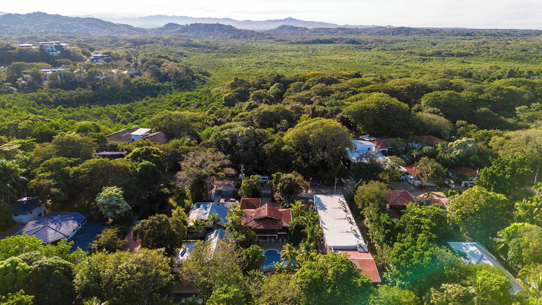 A residential area with houses surrounded by dense greenery and hills in the background.