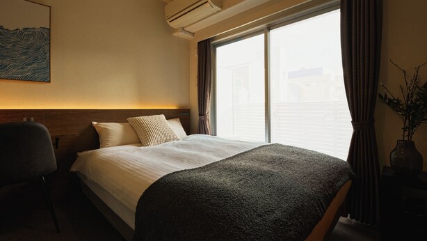 Una habitación de hotel con cama, escritorio con lámpara, silla, ventana con cortinas y aire acondicionado instalado en la pared.