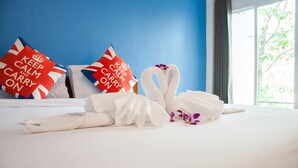 Un lit bien fait, avec du linge de lit blanc, deux oreillers à motifs rouges et blancs, et une serviette blanche ornée d’une fleur violette.