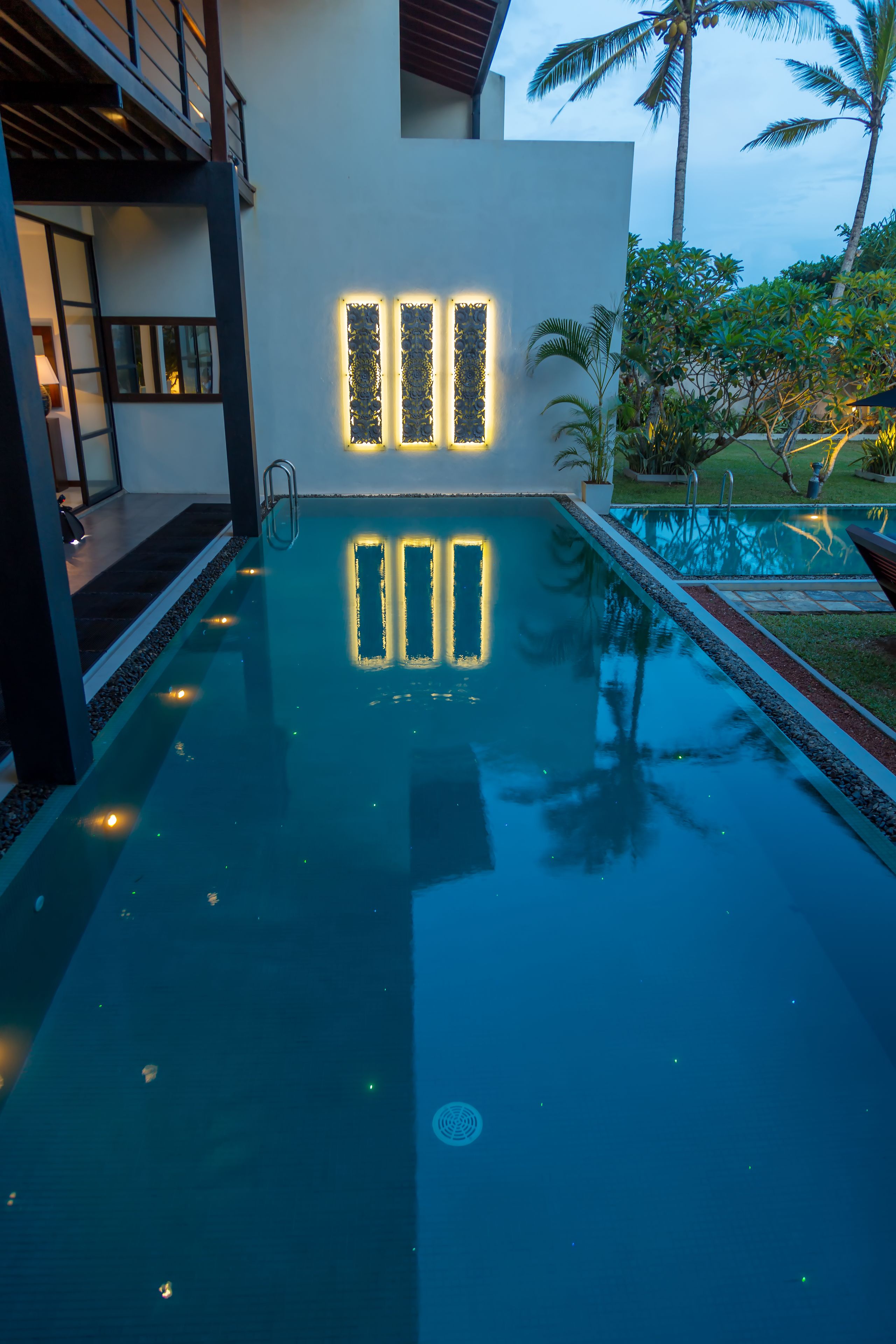 A swimming pool with illuminated panels, a modern building with large windows, and palm trees in the background.