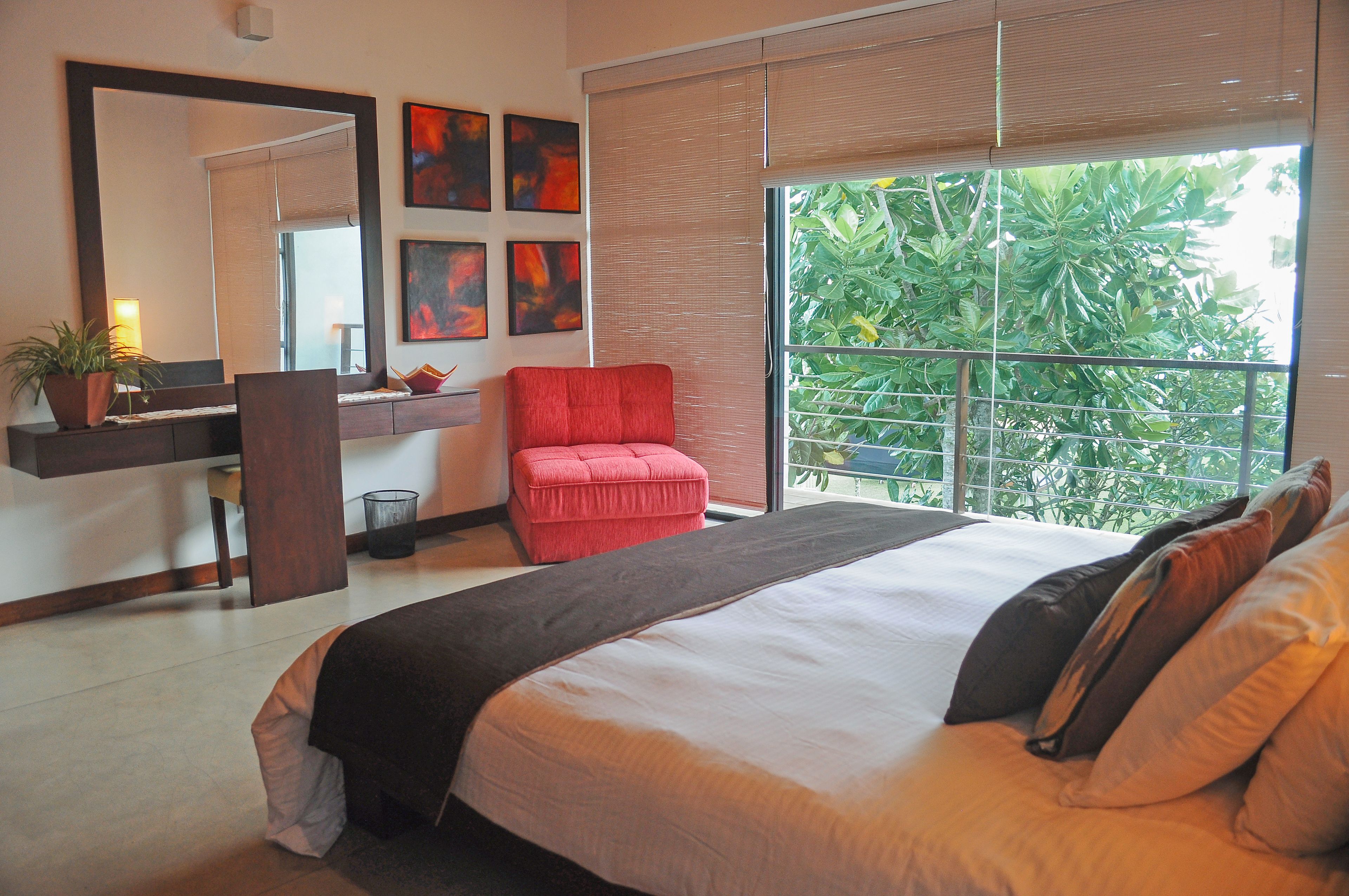 A bedroom with a large bed, a red chair, a desk with a mirror, and a window with a view of greenery.