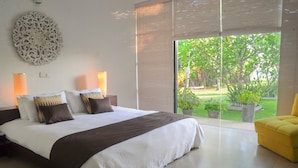 A modern bedroom with a large bed, two bedside lamps, a decorative wall piece, and a view of a garden through a sliding door. - Mirissa Beach Villa (Thalaramba)