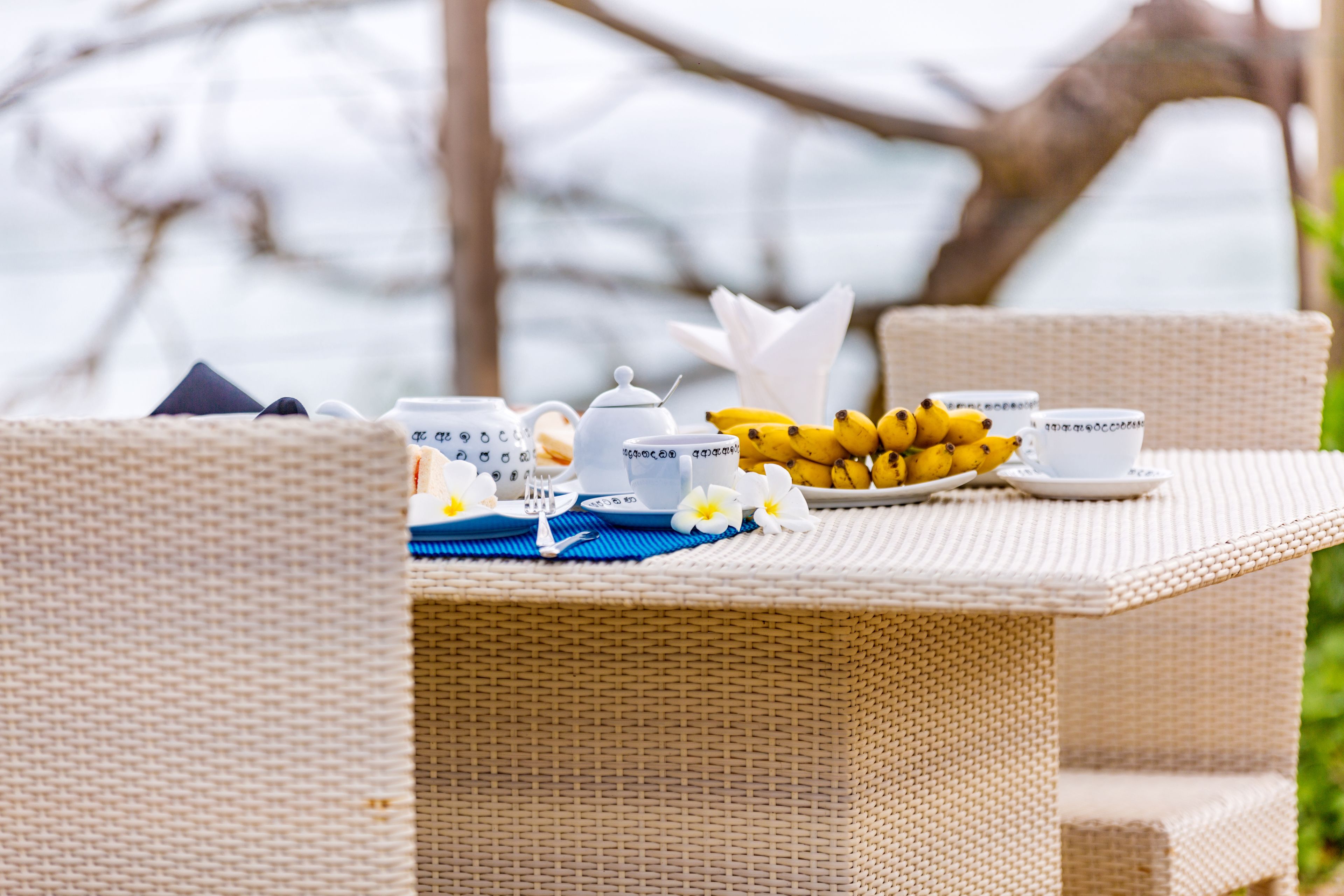 A wicker table set with tea cups, a teapot, a plate of bananas, and a blue placemat.