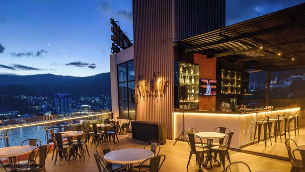 Une terrasse extérieure avec des tables et des chaises, un espace bar offrant une vue sur la ville, et une piscine en arrière-plan.