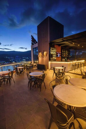 Een terras buiten met tafels en stoelen, een bar met uitzicht op de stad en een zwembad op de achtergrond.