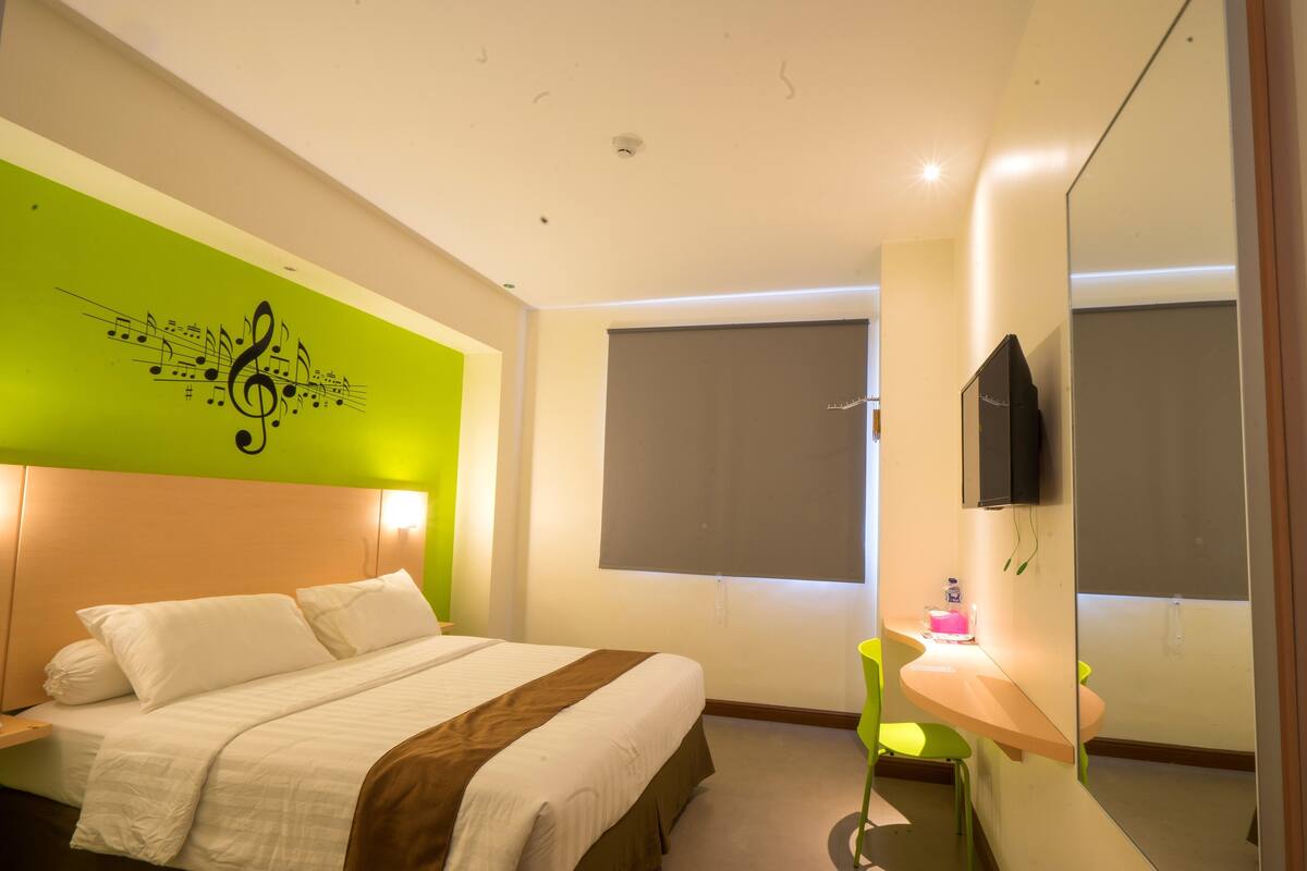 A hotel room with a bed, a green wall with a musical note design, a window with a blind, a TV mounted on the wall, and a small table with a pink object.