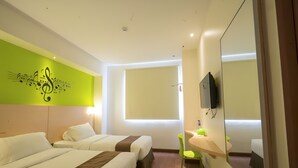 Une chambre d’hôtel avec deux lits, un mur vert orné d’un motif représentant une note de musique, une fenêtre, un miroir, une petite table avec une chaise verte et un téléviseur fixé au mur.