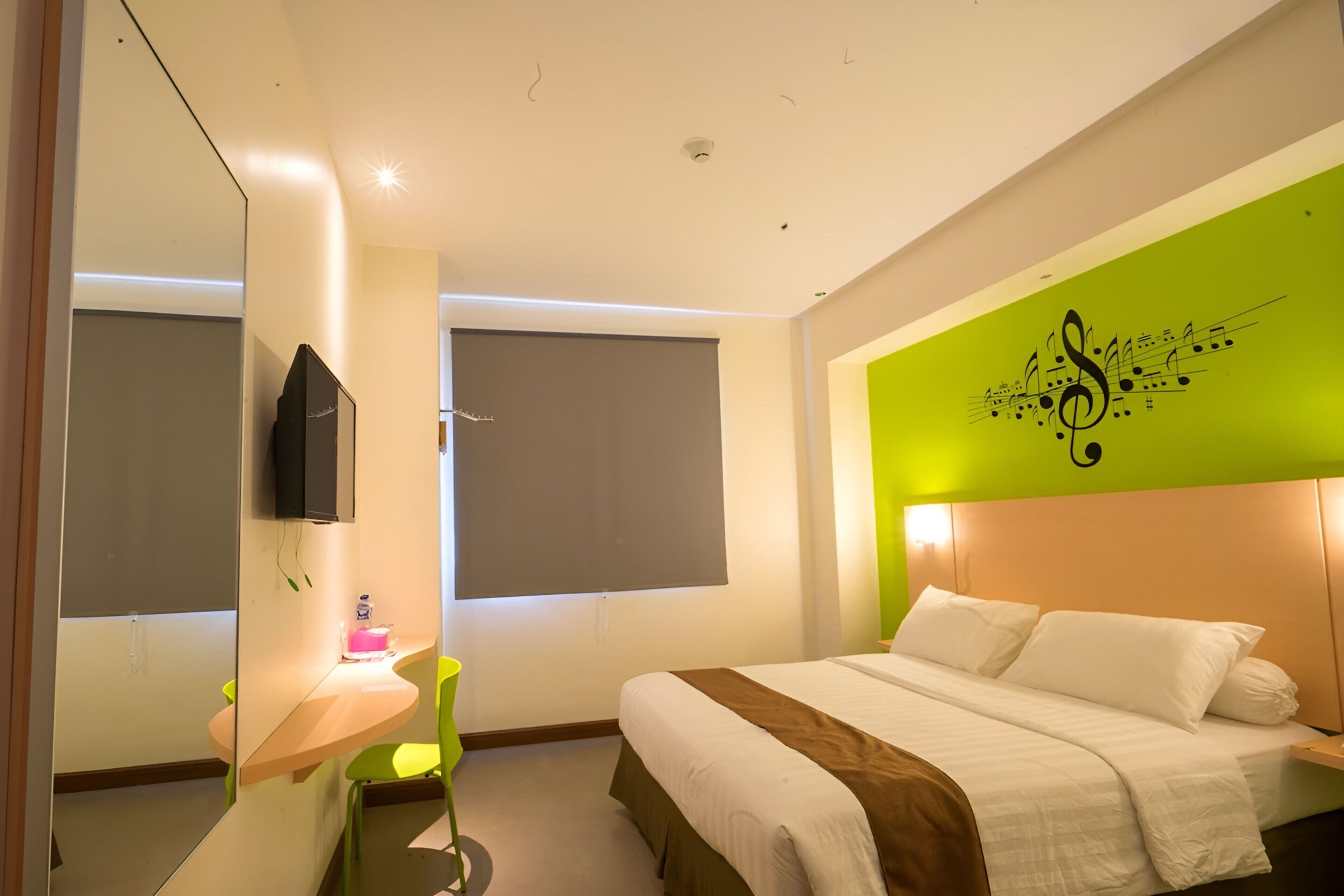 A modern hotel room with a bed, a TV, a desk with a chair, and a green accent wall.