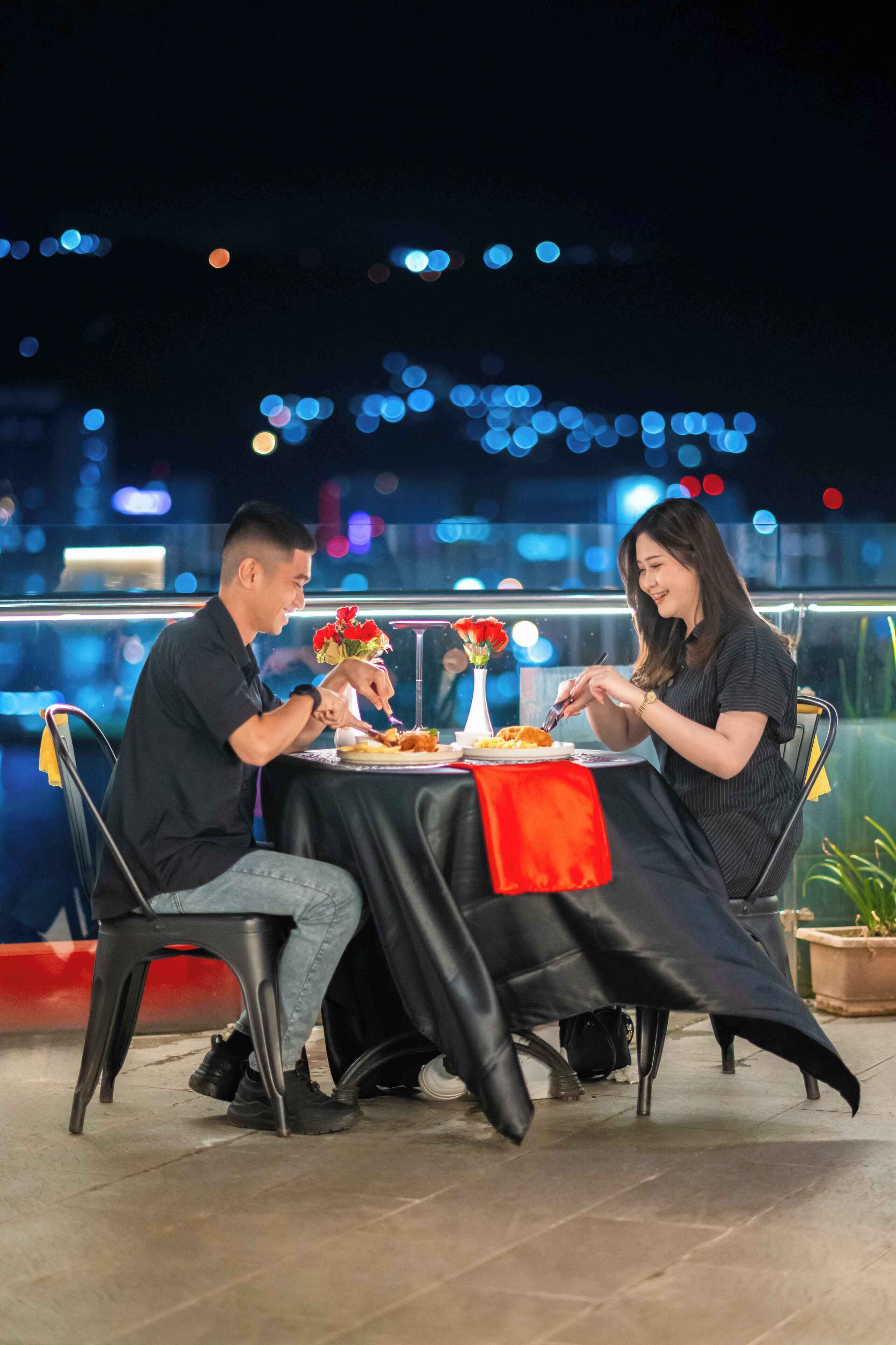 Dos personas cenando en una mesa al aire libre con vistas a la ciudad, un florero y un mantel rojo.