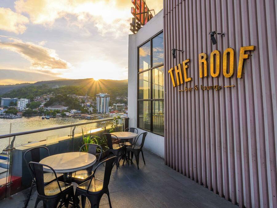 Un bar en la azotea con mesas y sillas, con vistas a un paisaje urbano al atardecer.