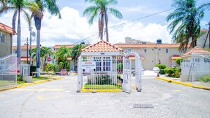 A gated entrance with a sign for visitors, palm trees, and buildings with red-tiled roofs.