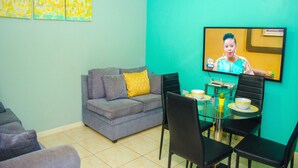 A living room with a grey sofa, a glass dining table with chairs, and a TV mounted on a teal wall.