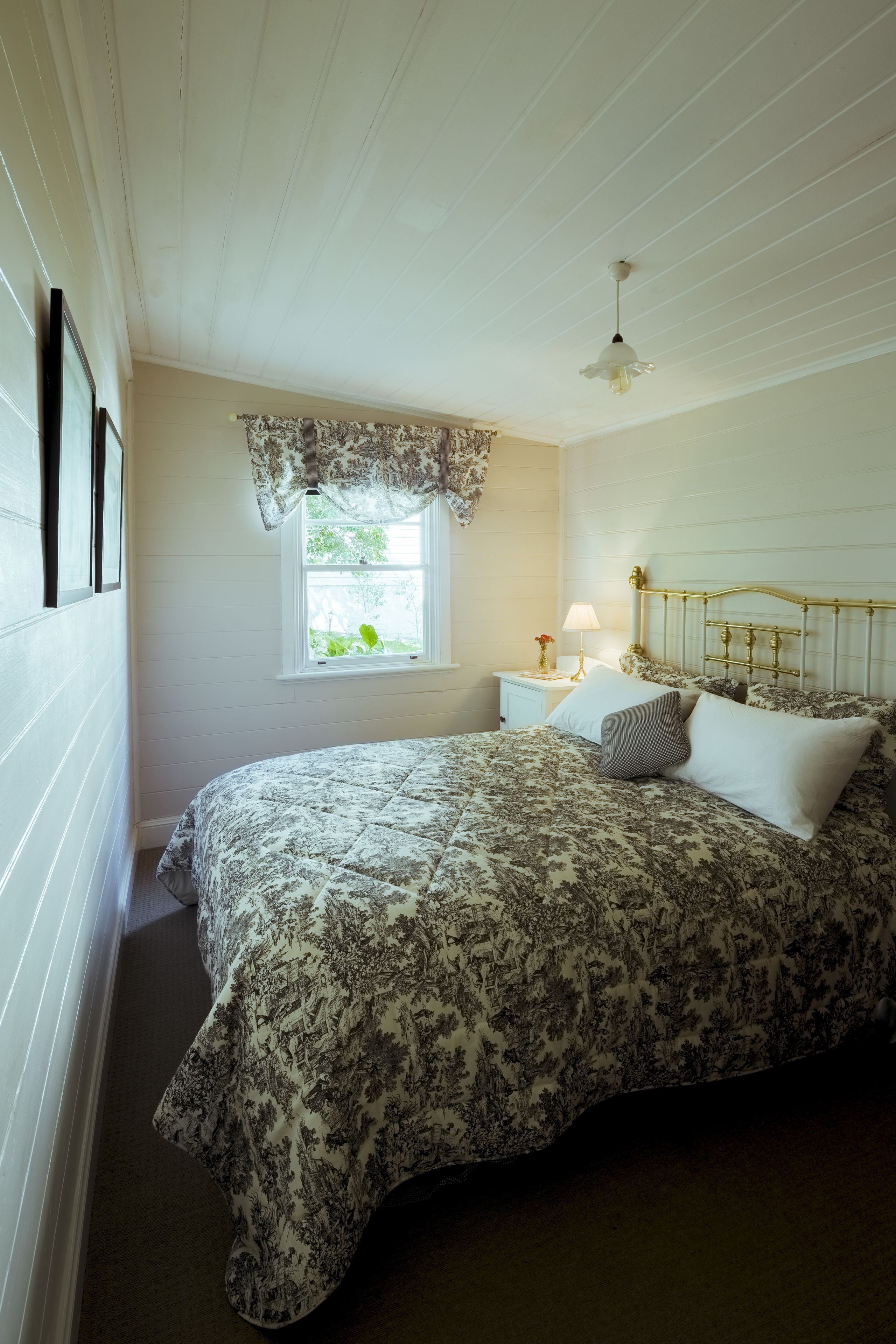 Une chambre à coucher comprenant un lit, une table de chevet, une lampe, une fenêtre donnant sur un paysage verdoyant et un ventilateur de plafond.