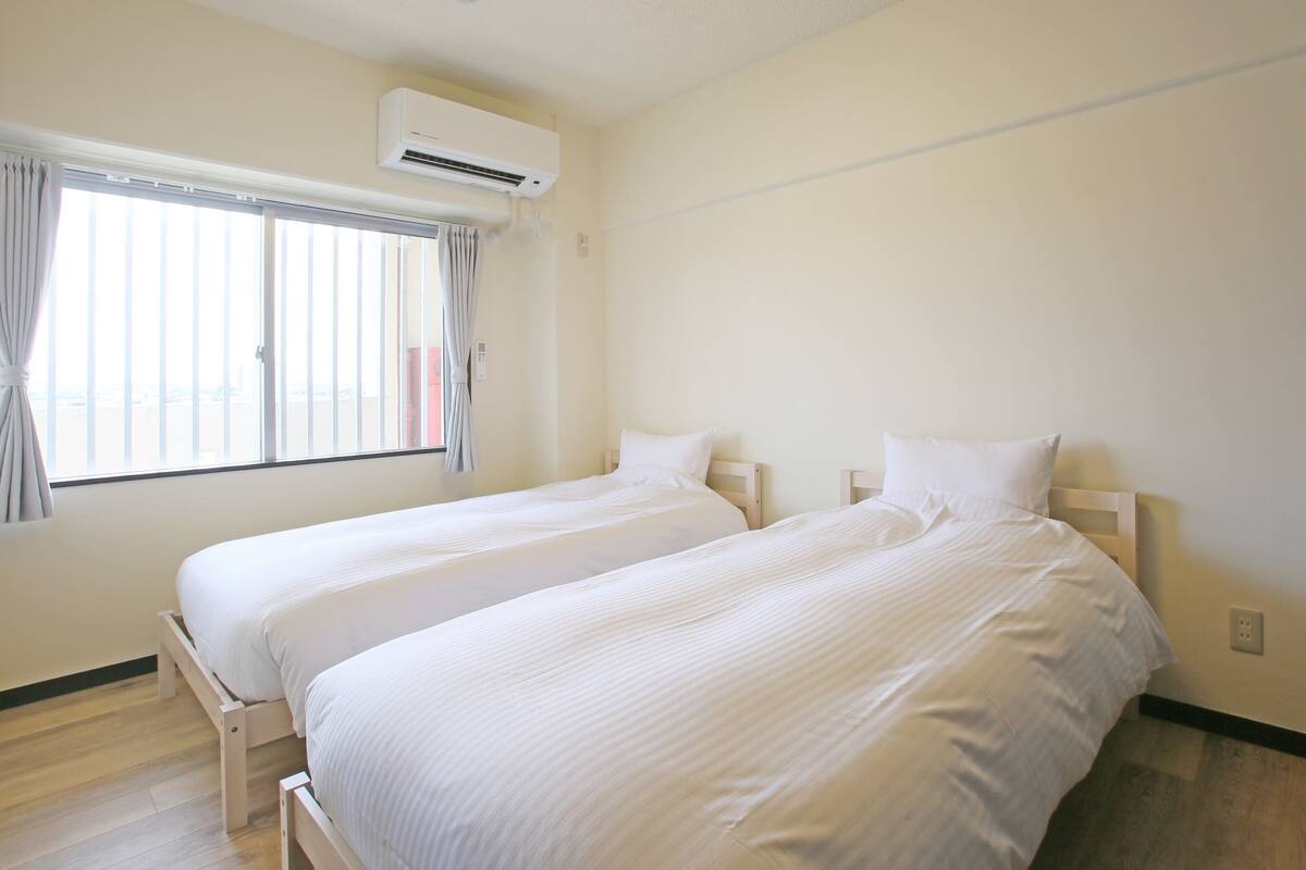 A hotel room with two beds, an air conditioning unit, a window with curtains, and a light fixture on the ceiling.