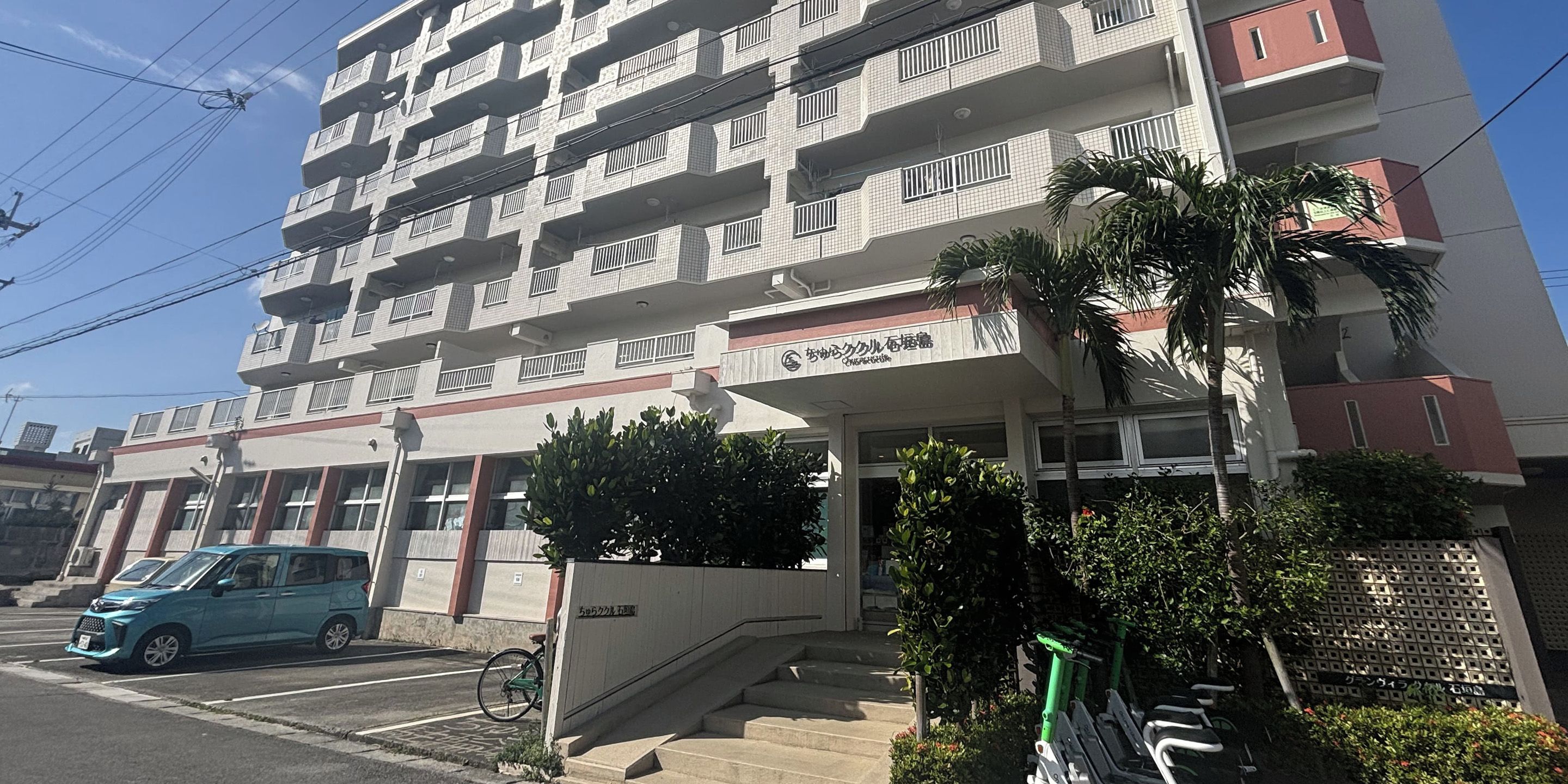 A multi-story residential building with balconies, a staircase leading to the entrance, and a parked car.