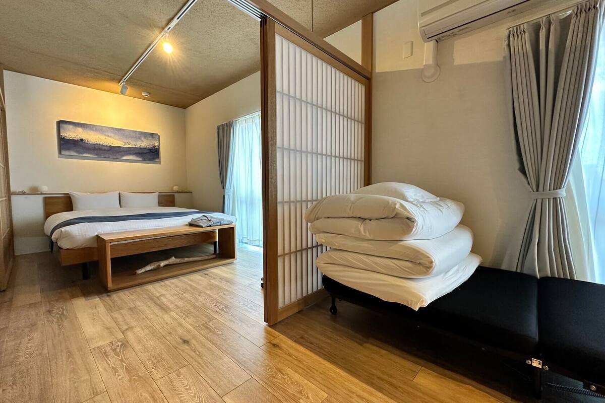 A modern hotel room with a wooden floor, a bed with white bedding, a black sofa, and a view of the outdoors through a window with curtains.