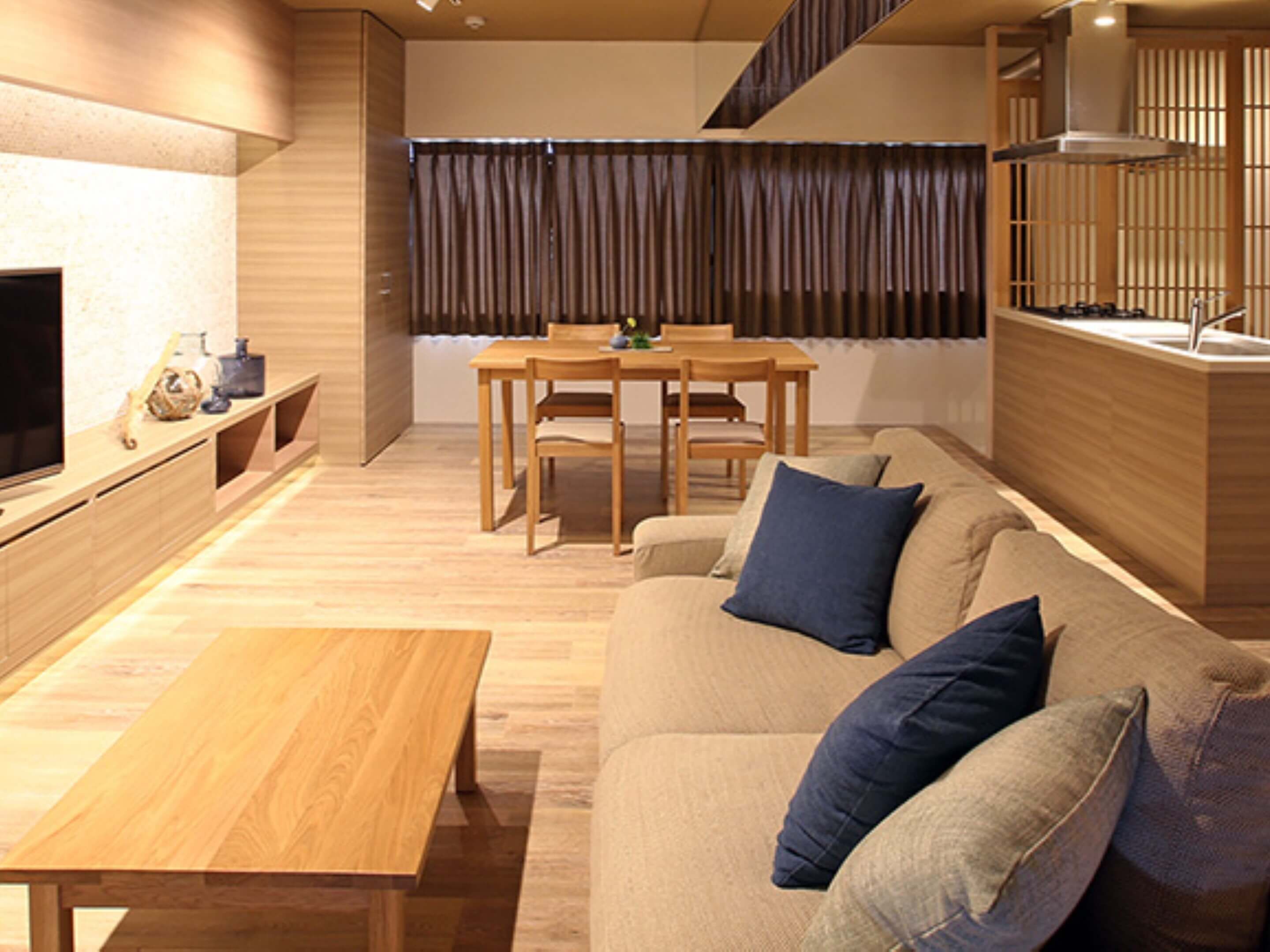 A modern living room with a wooden coffee table, a sofa with blue cushions, a dining area with a table and chairs, and a kitchen area with a stove.