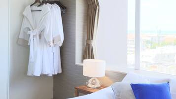 Una habitación de hotel con una cama, una mesita de noche con lámpara, una ventana con cortinas y un perchero con batas blancas.