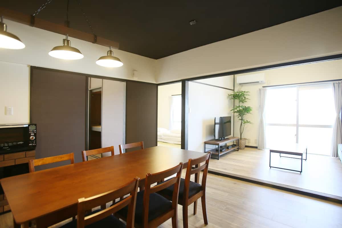 A modern dining area with a wooden table and chairs, a television on a stand, and a sliding glass door leading to a balcony.