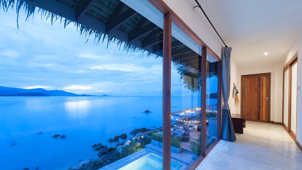 A modern hotel room with a large glass wall offering a view of a calm body of water and distant mountains. - Tamarina Bed & Beyond (Koh Samui)
