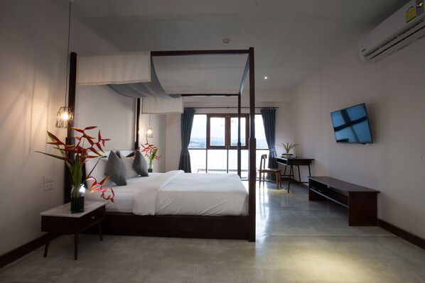 A modern hotel room with a four-poster bed, a desk with a chair, a TV mounted on the wall, and a large window with curtains.