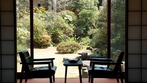 A room with tatami mat flooring, two black chairs, a small table, and a view of a garden with various plants and a rock. - Masuya Ryokan (Izumo)
