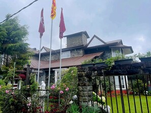 A stone building with a red roof, a flagpole with flags, and a garden with pink flowers. - The Rosedale Grand Nuwara Eliya (Nuwara Eliya)