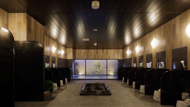 A modern, dimly lit restroom with wooden paneling, multiple sinks, and a central seating area with a view of trees through frosted glass. - Hotel Route Inn Ube (Ube)